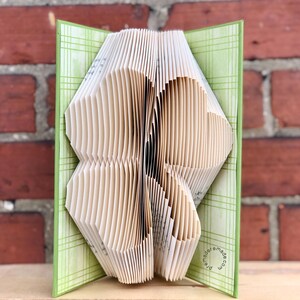 May include: A folded book sculpture with pages cut and shaped to form a heart design. The book's cover is light green with a plaid pattern. The background is a red brick wall. The book is open, revealing the heart shape.