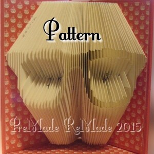 Könnte beinhalten: Eine gefaltete Buchkunstskulptur in Form eines Herzens. Das Buch besteht aus cremefarbenem Papier und hat einen roten Einband mit einem Muster aus kleinen Kronen. Das Buch trägt den Titel "Pattern" und am unteren Rand des Buches steht "Fred Made Rel Made 2015".