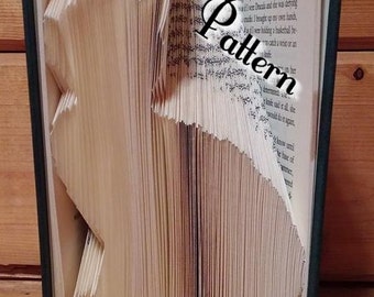 May include: A book folded into the shape of a cat. The book is open and the pages are folded to create the shape of the cat. The book is sitting on a wooden surface. The text "Pattern" is written in large letters on the book.