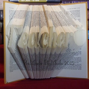 May include: A book with the word "Remarkable" folded into the pages. The book is open and the pages are folded to create the word. The book is on a red surface.