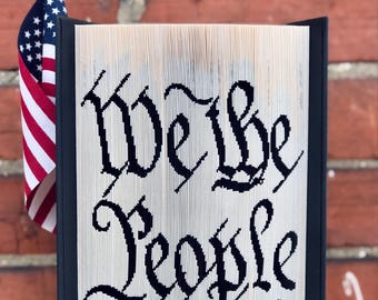 May include: A folded book art piece with the words "We the People" in black calligraphy. An American flag stands to the left of the book, with a red brick wall in the background. The book has a black frame.