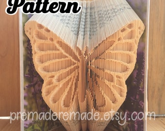 May include: A book folded into the shape of a butterfly. The butterfly is made from the pages of the book and is a light brown color. The butterfly is sitting on a wooden surface. The background is a blurry image of purple flowers.