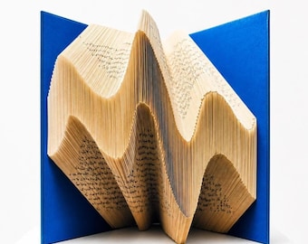 May include: A book with folded pages creating a three-dimensional wave pattern, featuring a bright blue hard cover. The pages are cream-colored with visible black printed text. The text "premaderemade.com" is printed at the bottom center.