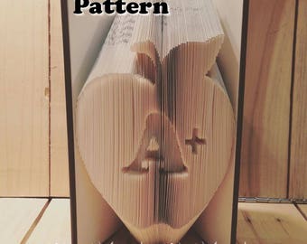 May include: A book folded into the shape of an apple with the letter A and a plus sign inside. The book is open and the text "Pattern" is visible on the front cover. The text "Premade Ready" is visible on the bottom of the book.
