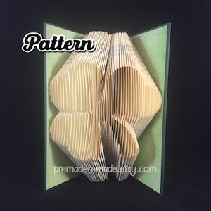 May include: A book art sculpture in the shape of a four-leaf clover. The book is folded and layered to create a 3D effect. The sculpture is displayed on a green background.