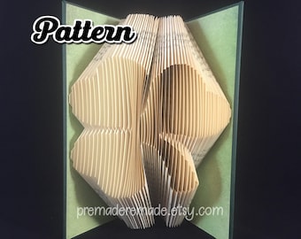 May include: A book art sculpture in the shape of a four-leaf clover. The book is folded and layered to create a 3D effect. The sculpture is displayed on a green background.
