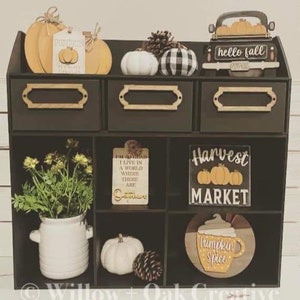 Puede incluir: Una estanter&iacute;a de madera negra con nueve compartimentos, decorada con art&iacute;culos de tem&aacute;tica oto&ntilde;al, incluyendo calabazas, calabacines, pi&ntilde;as y letreros que dicen "hello fall", "pumpkin patch", "I'm so glad I live in a world where there are Octobers", "Harvest Market", y "Pumpkin Spice".