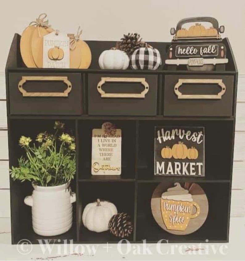 Puede incluir: Una estanter&iacute;a de madera negra con nueve compartimentos, decorada con art&iacute;culos de tem&aacute;tica oto&ntilde;al, incluyendo calabazas, calabacines, pi&ntilde;as y letreros que dicen "hello fall", "pumpkin patch", "I'm so glad I live in a world where there are Octobers", "Harvest Market", y "Pumpkin Spice".