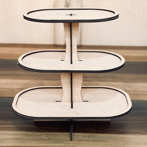 May include: Three-tiered wooden cake stand with a natural wood finish and black trim. Each tier is oval shaped.