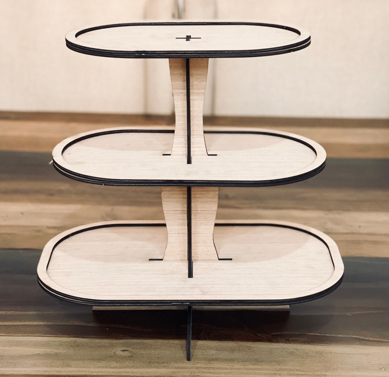 May include: Three-tiered wooden cake stand with a natural wood finish and black trim. Each tier is oval shaped.