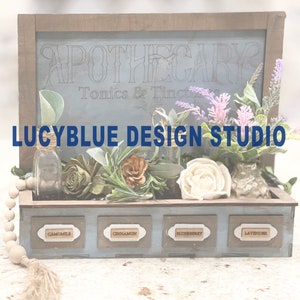 May include: A wooden apothecary box with a blue sign that reads "Apothecary Tonics & Tinctures" and "LUCYBLUE DESIGN STUDIO". The box has four drawers labeled "Chamomile", "Cinnamon", "Elderberry", and "Lavender".