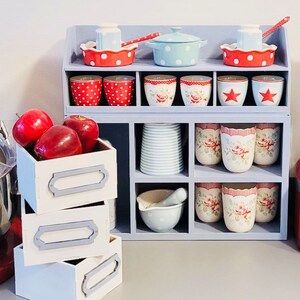 Puede incluir: Una estanter&iacute;a de madera azul con m&uacute;ltiples compartimentos, que contiene una colecci&oacute;n de tazas de lunares rojos y blancos, tazas rayadas azules y blancas y una fuente para hornear de patr&oacute;n azul y blanco. La estanter&iacute;a tambi&eacute;n tiene tres cajones blancos con ribetes grises.