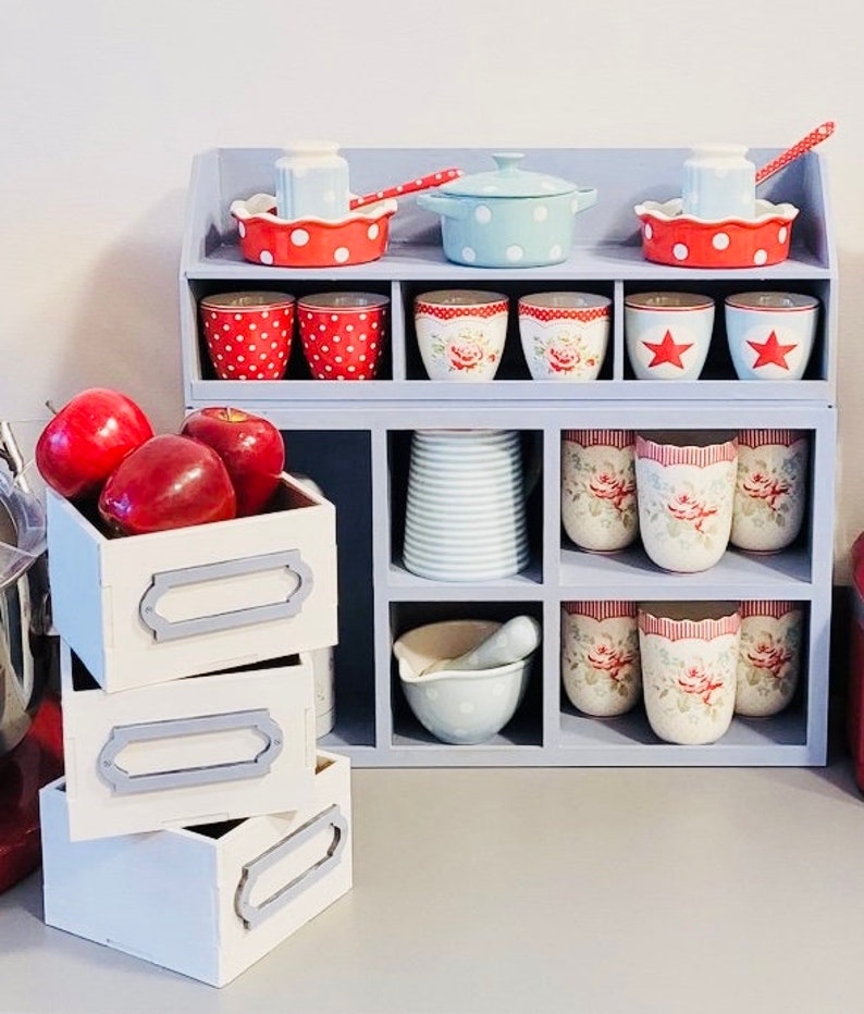 Puede incluir: Una estanter&iacute;a de madera azul con m&uacute;ltiples compartimentos, que contiene una colecci&oacute;n de tazas de lunares rojos y blancos, tazas rayadas azules y blancas y una fuente para hornear de patr&oacute;n azul y blanco. La estanter&iacute;a tambi&eacute;n tiene tres cajones blancos con ribetes grises.