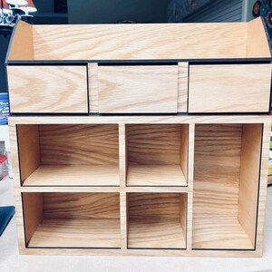 Puede incluir: Un organizador de madera con tres cajones y seis estantes. El organizador est&aacute; hecho de madera clara con ribetes negros.