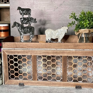 Puede incluir: Una caja de almacenamiento de estilo farmhouse de madera con un frente de malla de alambre. La caja tiene tres compartimentos y un estante superior. El estante está decorado con figuritas de animales de metal negro, una pequeña planta en maceta y un frasco de vidrio.