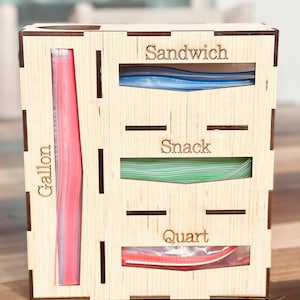 May include: Wooden organizer with four compartments for storing different sizes of plastic bags. The compartments are labeled "Gallon", "Quart", "Snack", and "Sandwich".