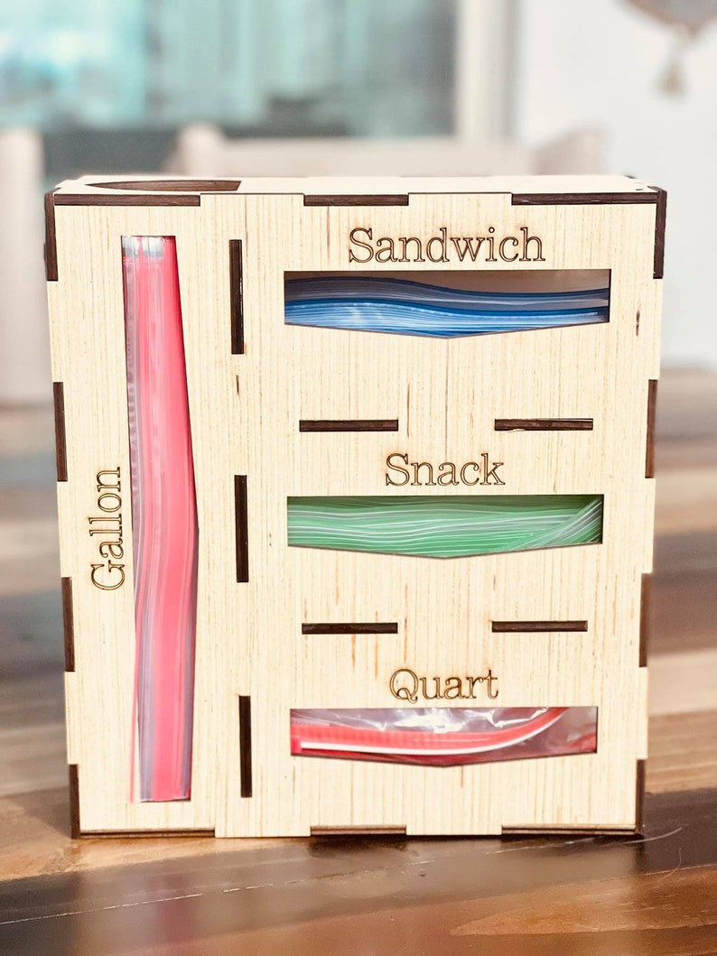 May include: Wooden organizer with four compartments for storing different sizes of plastic bags. The compartments are labeled "Gallon", "Quart", "Snack", and "Sandwich".
