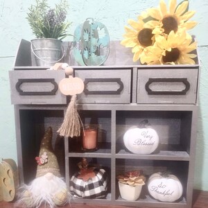 Puede incluir: Una estanter&iacute;a de madera gris de tres niveles con seis compartimentos. La repisa superior tiene tres cajones con ribetes negros. La repisa inferior tiene seis compartimentos. Los compartimentos est&aacute;n decorados con art&iacute;culos de tem&aacute;tica oto&ntilde;al, como calabazas, calabacines y girasoles. Los cajones est&aacute;n etiquetados como "Blessed", "Very Blessed" y "So Thankful".