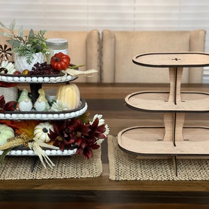 May include: A three-tiered wooden cake stand with a natural wood finish. The stand is empty and ready to be decorated with treats or other items. The stand is on a brown wooden table with a woven placemat.