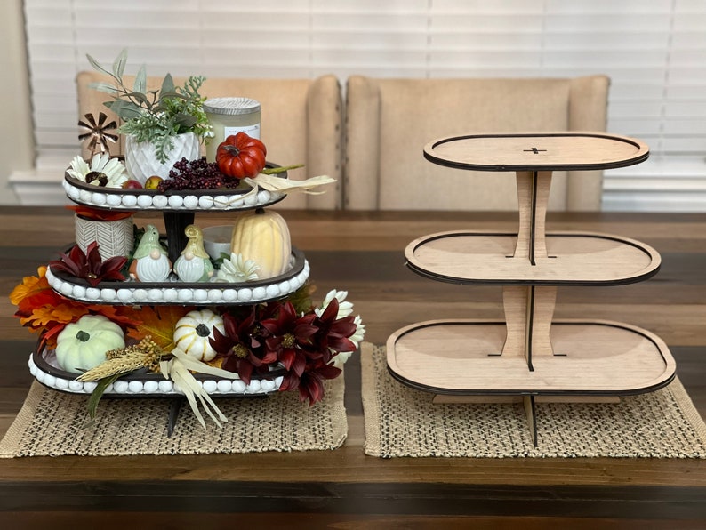 May include: A three-tiered wooden cake stand with a natural wood finish. The stand is empty and ready to be decorated with treats or other items. The stand is on a brown wooden table with a woven placemat.