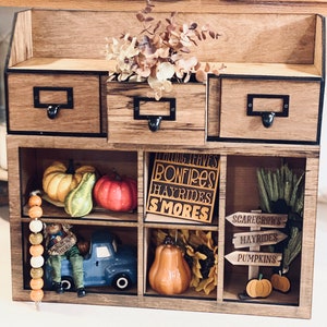 Puede incluir: Una estanter&iacute;a de madera con nueve compartimentos, decorada con diversos adornos de oto&ntilde;o. La fila superior tiene tres cajones con tiradores negros. La fila del medio tiene un cartel que dice "Falling Leaves, Bonfires, Hayrides, Smores" y otro que dice "Scarecrows, Hayrides, Pumpkins". La fila inferior tiene un cami&oacute;n de juguete, una calabaza y una guirnalda de cuentas.