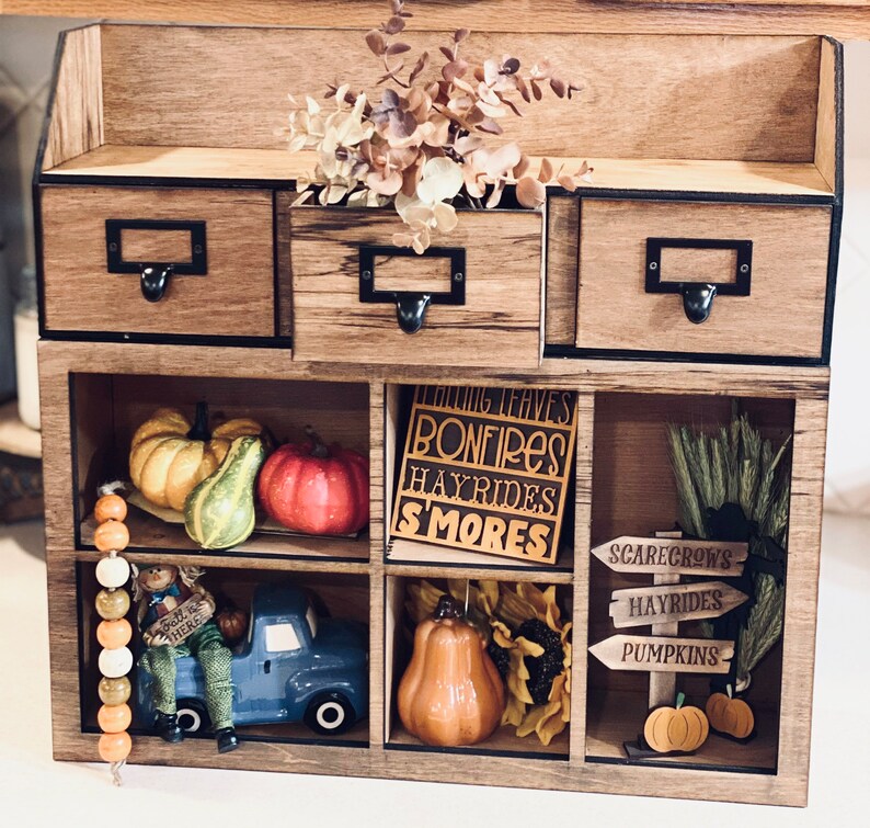 Puede incluir: Una estanter&iacute;a de madera con nueve compartimentos, decorada con diversos adornos de oto&ntilde;o. La fila superior tiene tres cajones con tiradores negros. La fila del medio tiene un cartel que dice "Falling Leaves, Bonfires, Hayrides, Smores" y otro que dice "Scarecrows, Hayrides, Pumpkins". La fila inferior tiene un cami&oacute;n de juguete, una calabaza y una guirnalda de cuentas.