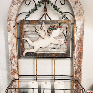 May include: A rustic wooden frame with a metal grate and a cutout of a cherub holding an arrow. Three white candles are displayed on a shelf below the frame.