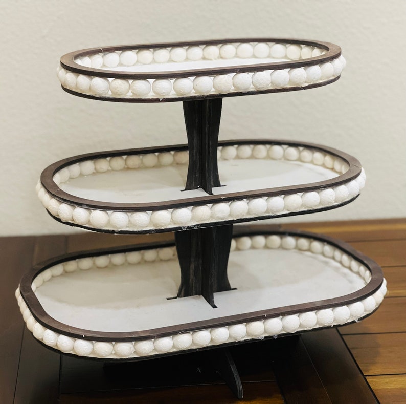May include: A three-tiered serving stand with a white oval base and white bead trim. The stand is made of wood and has a dark brown finish.