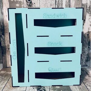 May include: A light blue wooden storage box with cutouts for storing various items. The box has labels for "Sandwich", "Snack", "Quart", and "Gallon".