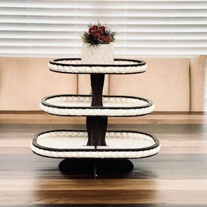 May include: A three-tiered white serving stand with a black base and white bead trim. The stand is oval shaped and is sitting on a wooden table.