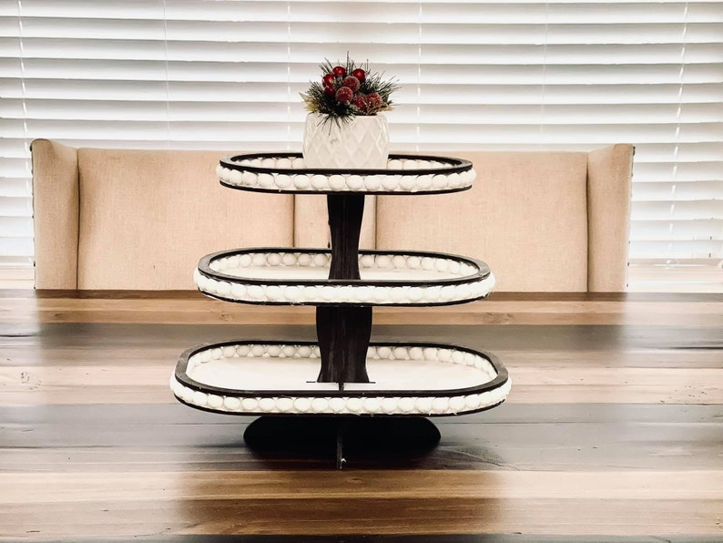 May include: A three-tiered white serving stand with a black base and white bead trim. The stand is oval shaped and is sitting on a wooden table.