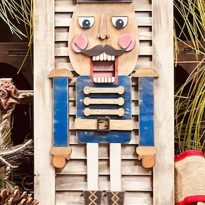 May include: A wooden nutcracker figurine with a blue hat, white beard, and brown boots. The nutcracker is painted with a whimsical expression and is standing on a wooden shutter.