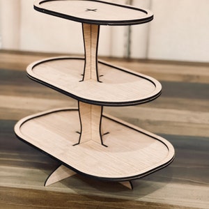 May include: A three-tiered wooden cake stand with a natural wood finish and black trim. Each tier is oval shaped and the stand has a wide base.