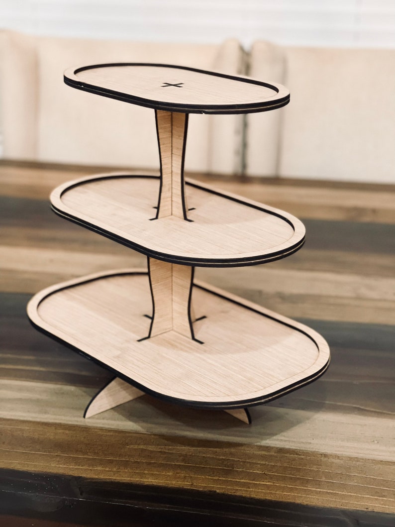 May include: A three-tiered wooden cake stand with a natural wood finish and black trim. Each tier is oval shaped and the stand has a wide base.