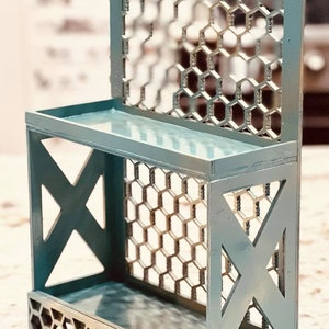 Op de afbeelding: Een tealblauw houten plank met twee planken en een honingraatpatroon aan de achterkant. De plank heeft een decoratief X-vormig ontwerp aan de zijkanten.