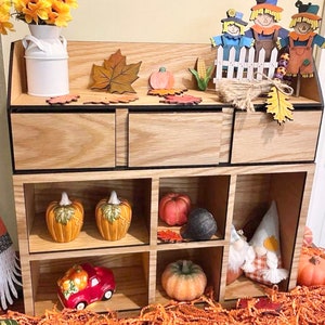 Puede incluir: Una estanter&iacute;a de madera con seis compartimentos, decorada con art&iacute;culos de tem&aacute;tica oto&ntilde;al, como calabazas, calabacines, espantap&aacute;jaros y hojas de oto&ntilde;o.