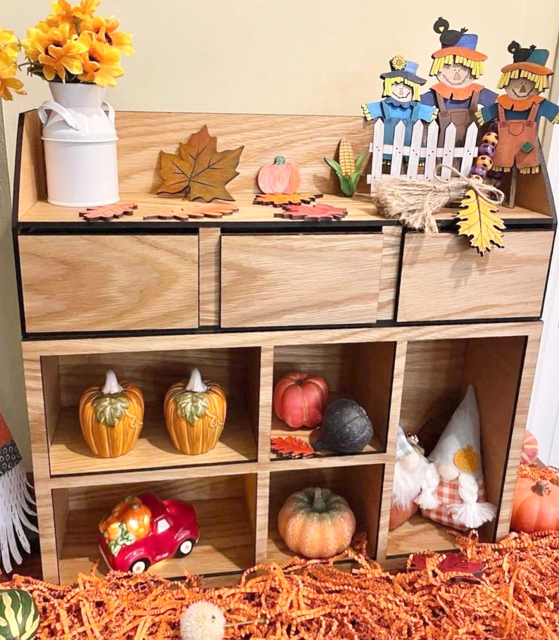 Puede incluir: Una estanter&iacute;a de madera con seis compartimentos, decorada con art&iacute;culos de tem&aacute;tica oto&ntilde;al, como calabazas, calabacines, espantap&aacute;jaros y hojas de oto&ntilde;o.