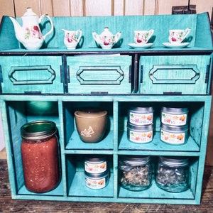Puede incluir: Una estanter&iacute;a de madera de color verde azulado con seis compartimentos. La repisa superior tiene cuatro peque&ntilde;as tazas de t&eacute; de porcelana y platillos. La repisa del medio tiene un taz&oacute;n de cer&aacute;mica marr&oacute;n y tres peque&ntilde;as latas de metal con etiquetas. La repisa inferior tiene un frasco de vidrio con tapa verde, un frasco de vidrio marr&oacute;n con tapa verde y dos frascos de vidrio transparente con tapas plateadas.