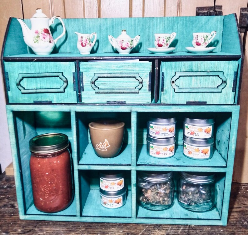 Puede incluir: Una estanter&iacute;a de madera de color verde azulado con seis compartimentos. La repisa superior tiene cuatro peque&ntilde;as tazas de t&eacute; de porcelana y platillos. La repisa del medio tiene un taz&oacute;n de cer&aacute;mica marr&oacute;n y tres peque&ntilde;as latas de metal con etiquetas. La repisa inferior tiene un frasco de vidrio con tapa verde, un frasco de vidrio marr&oacute;n con tapa verde y dos frascos de vidrio transparente con tapas plateadas.