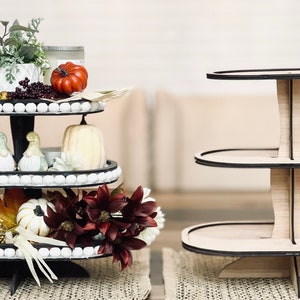 May include: A three-tiered wooden serving tray with black trim. The top tier is decorated with fall-themed items, including a small potted plant, a pumpkin, and a small windmill. The bottom two tiers are empty.
