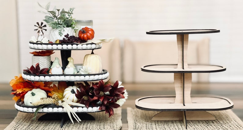 May include: A three-tiered wooden serving tray with black trim. The top tier is decorated with fall-themed items, including a small potted plant, a pumpkin, and a small windmill. The bottom two tiers are empty.