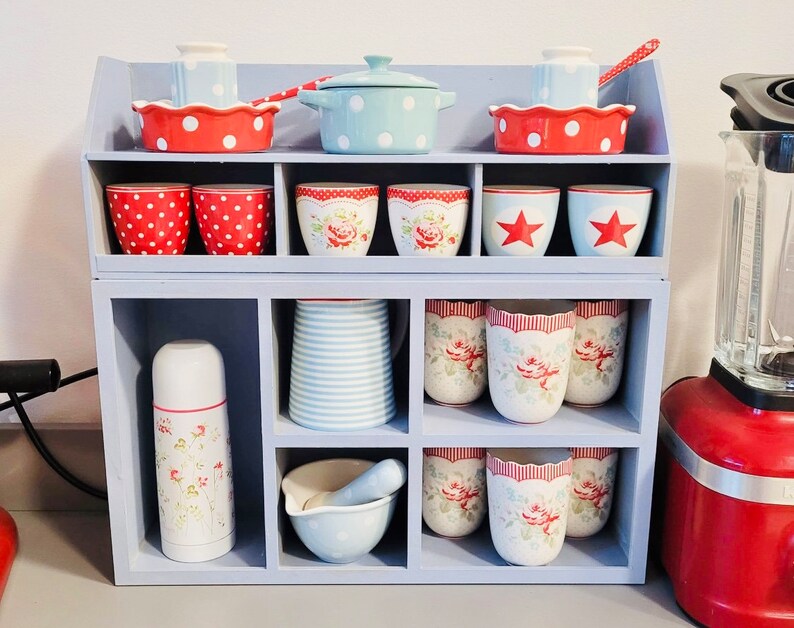 Puede incluir: Una estanter&iacute;a blanca con una variedad de tazas, cuencos y una tetera con patrones rojos y blancos. La estanter&iacute;a est&aacute; llena de utensilios de cocina y est&aacute; colocada sobre una encimera.