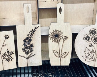 Puede incluir: Cuatro tablas de cortar de madera con diseños florales grabados con láser. Los diseños incluyen una amapola, un grupo de flores, una margarita y un grupo de flores más pequeñas.