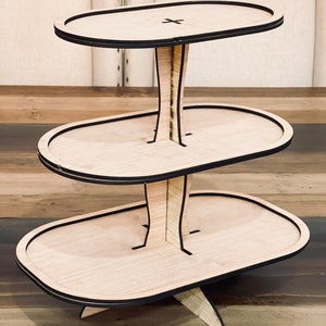 May include: Three-tiered wooden serving stand with oval-shaped trays. The stand is made of light brown wood with black trim around the edges of the trays. The stand is designed to hold desserts, snacks, or other small items.