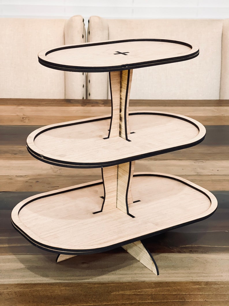 May include: Three-tiered wooden serving stand with oval-shaped trays. The stand is made of light brown wood with black trim around the edges of the trays. The stand is designed to hold desserts, snacks, or other small items.