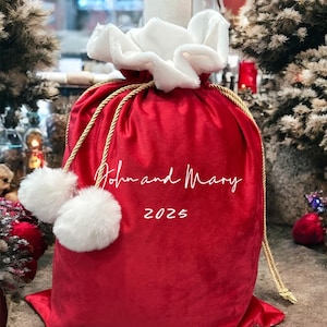 May include: A red velvet gift bag with a white fluffy trim and two white pom-poms. The bag has a gold drawstring and the text "John and Mary 2025" in white script. The bag is designed for gift-giving.