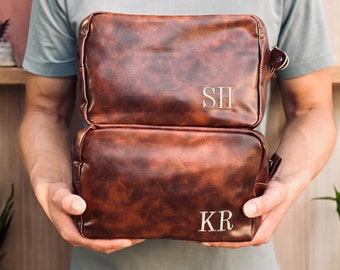 May include: Two brown leather travel bags with zipper closures. The top bag has the initials "SH" embroidered on the front. The bottom bag has the initials "KR" embroidered on the front.