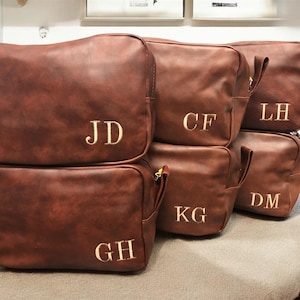 Op de afbeelding: Een stapel bruine leren toilettassen met goud geborduurde initialen. Elke tas heeft een handvat en een ritssluiting. De initialen die op de tassen zijn geborduurd zijn "JD", "CF", "LH", "KG", "DM" en "GH".