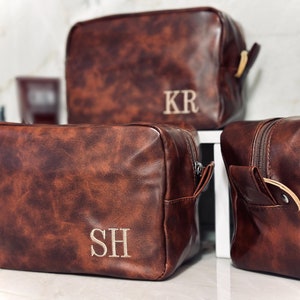 May include: Three brown leather toiletry bags with zipper closures. The bags have gold accents and are personalized with the initials "KR", "SH", and "JW".