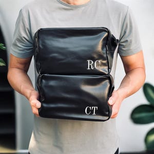 Op de afbeelding: Twee zwarte, rechthoekige tassen worden vastgehouden. De bovenste tas heeft de initialen "RC" in wit, en de onderste tas "CT". De tassen zijn gemaakt van een glad, leerachtig materiaal. Ideaal voor toiletartikelen of reizen.